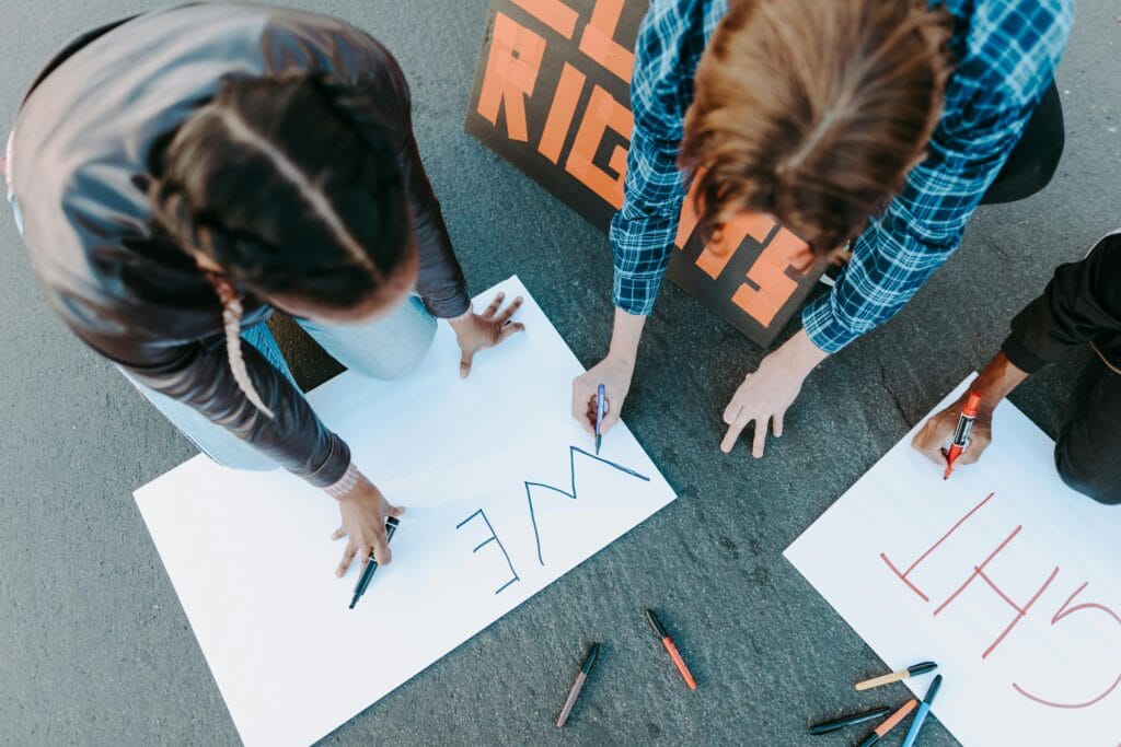 People writing protest signs on the pavement, seen from above. Outdoor daytime scene.